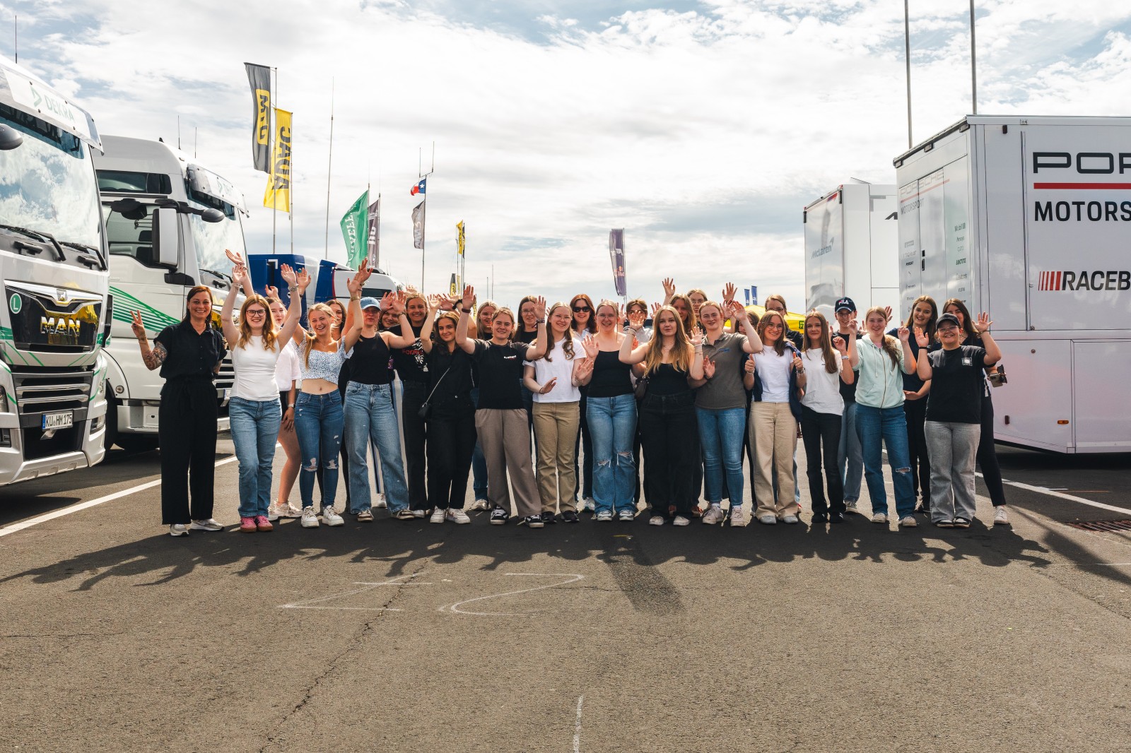 Gruppenfoto von Teilnehmerinnen der Discover Motorsport – Young Women’s Edition 2025 am Nürburgring mit erhobenen Händen vor den DTM-Teamtruckern, dazu das Event-Logo und der Hinweis „Am 23. Juli in Oschersleben – jetzt anmelden“.